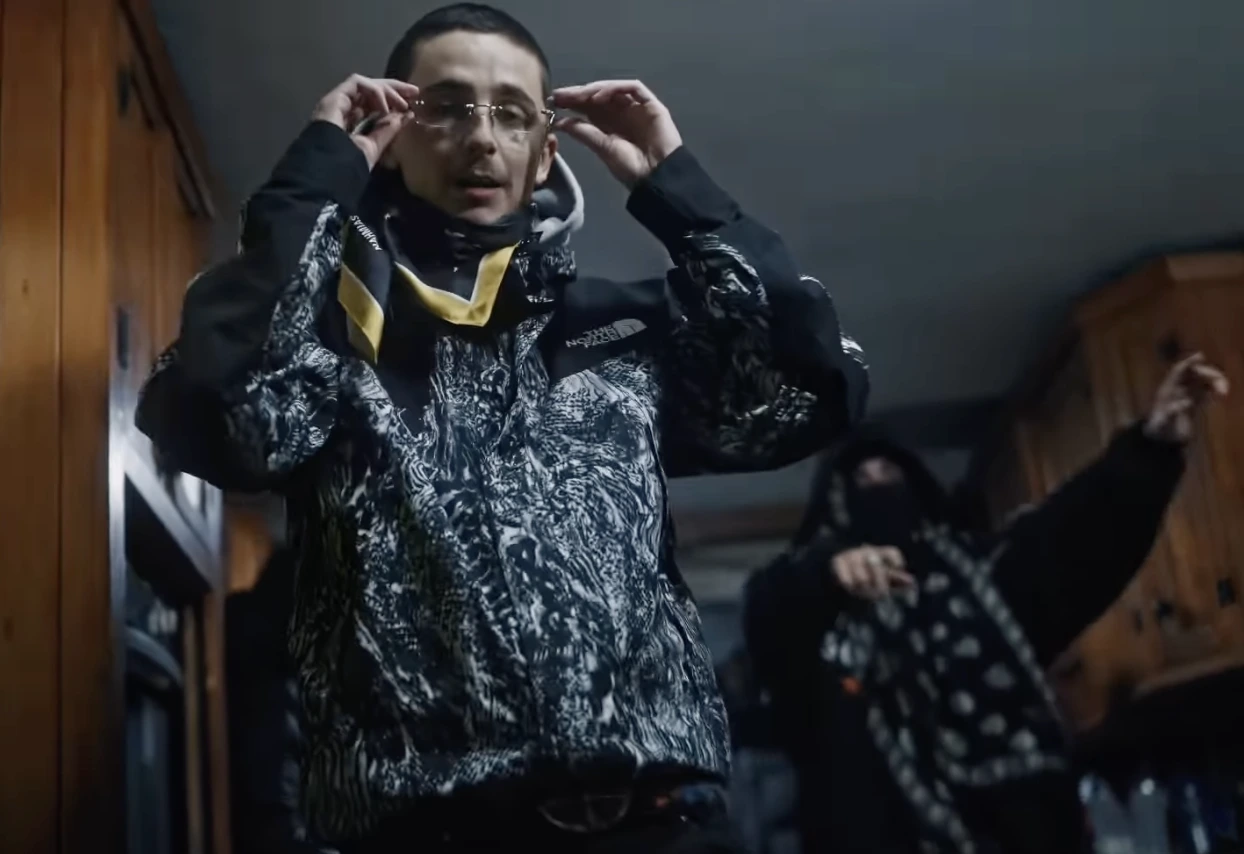 For weeks, the internet entertained a theory that felt deliberately unresolved. A masked British rapper gaining rapid attention. An Oscar nominated actor with a documented interest in hip hop. Subtle overlaps in timing, aesthetics, and attitude. The idea that Timothée Chalamet could be hiding behind the alias EsDeeKid became a recurring topic across social media and entertainment press. That speculation has now reached a clear conclusion. Timothée Chalamet appears directly alongside EsDeeKid on the remix of “4 Raws,” placing both figures in the same frame and effectively closing the question of identity. The collaboration confirms that Chalamet was not the anonymous rapper, but something arguably more interesting: a willing participant in the project itself. EsDeeKid emerged in 2025 as one of the UK rap scene’s most closely watched new names. Performing masked and avoiding public appearances, the artist built momentum through streaming platforms and short form video culture, with “4 Raws” becoming one of the most circulated tracks of the year. The anonymity invited curiosity, and the rumors linking the project to Chalamet escalated during the actor’s press appearances in London, where he avoided giving a direct answer when asked about the theory. The remix reframes the narrative. Chalamet does not parody rap culture, nor does he position himself as a novelty feature. His verse is delivered with restraint, referencing his current film work and public life without irony. The tone aligns with the track’s existing mood rather than disrupting it, suggesting a considered collaboration rather than a publicity stunt. For Chalamet, music is not unfamiliar territory. Long before his rise in cinema, he performed under the name Lil Timmy Tim during his teenage years, a detail that has periodically resurfaced in interviews. What makes this moment different is its timing and context. Appearing on a track that was already culturally active, rather than launching an independent project, positions him as a contributor rather than a crossover headline. For EsDeeKid, the collaboration expands visibility while preserving mystique. The rapper remains masked, unnamed, and separate from the celebrity machine, even while sharing a track with one of the most recognizable actors of his generation. In an era where anonymity is increasingly rare, the decision to keep that distance feels intentional and strategic. Rather than feeding speculation further, the release resolves it with clarity. Chalamet is not EsDeeKid. He is simply present, briefly and deliberately, inside the world the rapper created. The mystery ends not with a reveal, but with a collaboration that feels unexpectedly grounded.