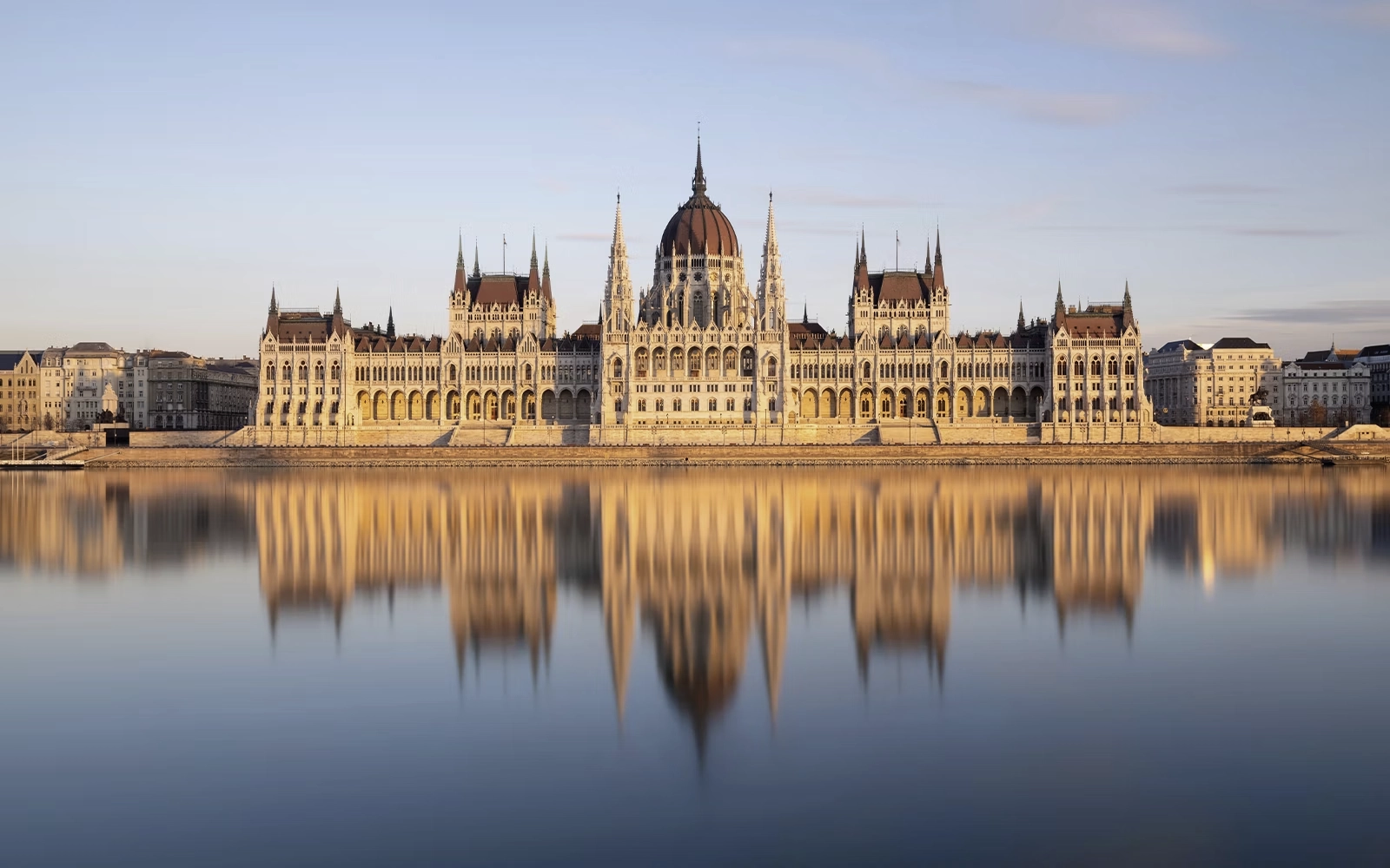 Hungarian Parliament Building, Budapest, Hungary On the banks of the Danube, the Hungarian Parliament Building presents an elaborate façade of pointed arches, slender spires and sculpted ornament beneath a commanding central dome. Designed by Imre Steindl, it was built between 1885 and 1904 in the Gothic Revival style. Inside, the ornate Grand Stairway leads to the Dome Hall, conceived as the architectural center of the building. The view from across the river reveals the full composition, with the dome rising between two symmetrical wings.